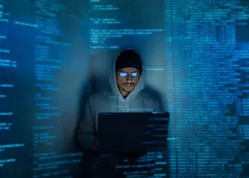 hooded young adult using laptop in dark room